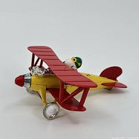 Side view of the Aviva Snoopy Red Baron toy plane showing yellow metal body, red wings, and sitting Snoopy pilot figure with green helmet and goggles.