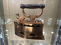 Front and full view of the copper color vintage charcoal iron, showing wooden handle, hinged top, and rooster ornament on corner.