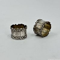 Two sterling silver napkin rings shown side by side on white background, highlighting the scroll pattern edges and cylindrical shape.