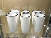 Six white milk glasses with black rims arranged in two rows of three on a glass shelf.