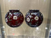 Front view of two round cranberry glass votive holders with hand-painted white daisies and blue flowers.