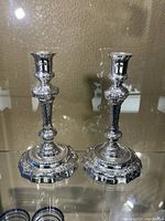 Pair of reflective metal candlestick holders shown side by side on glass surface, showcasing detailed ornate bases and stems.