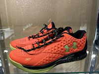 Side view showing bright orange Under Armour basketball shoe with green logo, black midsole with green speckles, and green outsole.