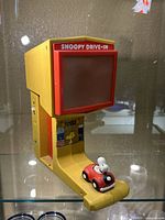 Front view of Snoopy Drive-In Movie Theater toy showing yellow body, red screen frame, Peanuts character graphics, and Snoopy in a red car on the track.