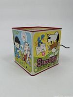 Side view showing Snoopy, Woodstock, Lucy, Linus, and Charlie Brown graphics on metal box with crank handle.
