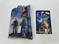 Photo showing X-Men Rogue action figure and E.T. powered bicycle toy in original blister packaging with visible wear and defects on packaging