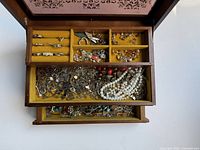 Top view of open wooden jewelry box showing multiple sections with assorted costume jewelry inside including rings and brooches.