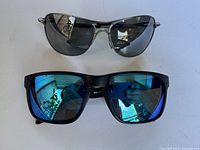 Two pairs of men's sunglasses. One pair is a silver metal frame rectangular shape with dark lenses. The other pair is a black plastic frame with blue mirrored lenses.