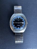 Photo of full Timex Marlin watch showing round case, metal expandable band, black dial with blue details, and date window.