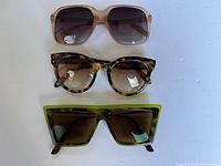 Three pairs of women's sunglasses arranged vertically on a white surface. Includes various frame shapes and colors: beige, tortoiseshell, and green.