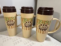 Photo of three identical beige Tim Hortons travel mugs with brown lids, showing front Tim Hortons Cafe & Bake Shop logos and tree branch leaf designs, placed on a table.