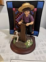 Figurine of a young Amish boy in a straw hat holding a wooden fence, standing with his dog on a garden base with flowers and a colorful mini quilt backdrop.