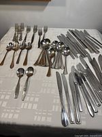 Full spread of assorted cutlery laid out on white patterned cloth, grouped by forks, spoons, knives, and butter spreaders.