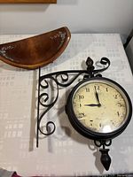 Photo showing Baribocraft wood bowl with floral painting and double-sided vintage replica wall clock with scroll wrought iron bracket on table.