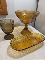 Four vintage glass pieces displayed on a white cloth: a large amber carnival glass fruit bowl with scalloped edge and grape pattern, a long oval carnival glass bread basket with diamond lattice texture, a smaller brown glass candy dish with circular pattern and footed base, all in excellent vintage condition.