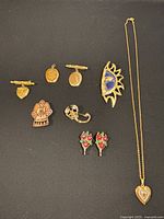 Overview of 7 golden-tone brooches, 1 pair of red rose earrings, and 1 gold-tone necklace with heart pendant on black background.