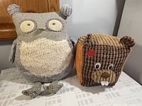 Two decorative stuffed animals on a table: a beige and gray owl and a corduroy-style weighted beaver with a red patch on its head.