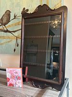 Glass front wooden display cabinet with dark finish and carved details on edges, showing shelves inside behind glass. Cabinet is standing on a gray table with colorful floral canvas art beside it.