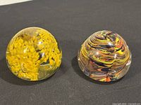 Photo showing both glass paperweights side by side on black surface, highlighting size and color contrast.