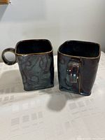 Pair of handmade square pottery coffee mugs shown side-by-side with visible brown and black iridescent glaze and subtle swirl patterns.