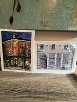 Photo showing two prints: one of a brick building Classic Video and the other a stone building Coffee and Company, both with artist sketches on the back of the matte.