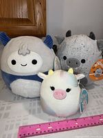 Three new with tags Squishmallows plush toys: Candess the Cow, Horace the Yeti, and Gio the Gargoyle arranged on a surface.