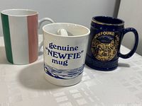 Three vintage Newfoundland-themed mugs displayed on a table: a white 'genuine NEWFIE mug', a dark blue Newfoundland crest mug, and a tricolor striped mug.