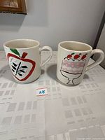 Two white ceramic mugs shown front view on table. One mug has an apple painted on side, other displays a cake with cherries.