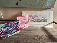Three art pieces displayed on wooden surface including a purple and teal abstract, a floral piece, and a framed print wrapped in plastic.