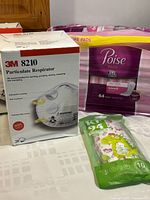 Photo showing one box of 3M 8210 particulate respirator masks, one pack of Poise bladder support pads with 64 count, and one pack of 10 kids face masks in animal prints.