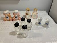 Five pairs of salt and pepper shakers displayed on a white surface. Includes ceramic bear chefs, clear glass shakers with black tops, and white porcelain shakers with blue floral design.