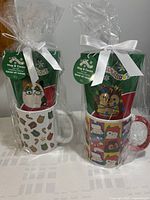 View of two Squishmallow-themed mugs each wrapped as gift sets with hot cocoa packets and tied with white ribbons.
