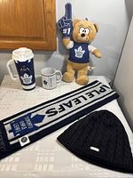 Photo showing all lot items on table: teddy bear wearing Toronto Maple Leafs shirt and holding foam finger, travel mug, ceramic mug, long scarf, and black knitted toque.