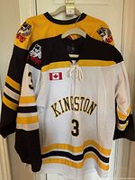 Both jerseys hanging, showing front details including lace-up neck, Kingston logo, number 3, Canadian flag, and wolf logo on sleeves.
