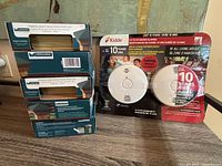 Photo of boxed two-pack Kidde smoke/fire alarms with original packaging, alongside five boxes of Veranda fence post caps stacked