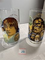 Two clear collectible drinking glasses featuring colorful 1977 Star Wars character graphics including Luke Skywalker, Princess Leia, Chewbacca, Han Solo and others.
