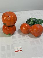 Pair of vintage ceramic orange tomato salt and pepper shakers stacked vertically next to orange radish-shaped salt and pepper shakers with green leafy tops, placed on a white cloth surface.