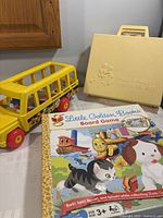 Photo showing the yellow Fisher-Price School Bus toy, cream-colored Fisher-Price Medical Kit case, and Little Golden Books board game box on a table.