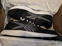 Side view of black Reebok sneaker showing white logo, white midsole, black outsole, and mesh upper inside original box.