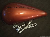 Photo of a metallic orange Harley-Davidson gas tank with black logo, attached electrical cord and switch, displayed on carpet.