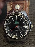 Close-up front view of Lacoste men's watch face showing black dial, silver indices, red seconds hand, Lacoste logo, tachymeter bezel, and water resistance marking.