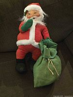 Full view of vintage Annalee Santa Claus doll standing with green sack at side on dark fabric background.