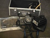 Photo showing the Olympus OM-2S Program camera with attached lens, flash unit, camera strap, manual in plastic sleeve, and hard carrying case.