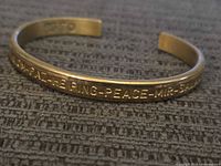 Close-up front view of gold-tone open cuff bracelet with engraved peace-related words.