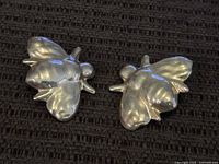 Pair of silver fly or bee-shaped earrings on dark fabric background.