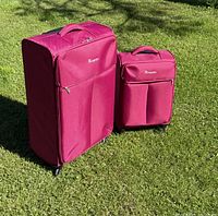 Two pink soft-side suitcases, one large and one carry-on size, standing on grass with handles retracted.