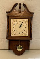 Front view of the antique wooden grandmother style wall clock showing the clock face with floral designs and brass pendulum below.
