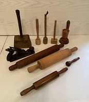 Photo showing nine antique wooden tools arranged on a wooden surface against a wood-paneled backdrop. Items include hand plane, mallets, rolling pins, pestle, and hooked tool.