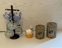 Photo showing six clear glass mugs with blue goose design on a dark wood stand, white ceramic goose figurine on basket base, and two vintage goose-themed metal tins.