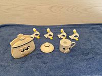 Top-down view of a blue cloth background with four ceramic goose napkin holders, a lidded ceramic container with goose decoration, an extra lid, and a mug with a goose design.